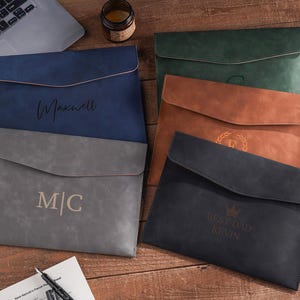 Puede incluir: Varios portadocumentos de cuero personalizados en azul marino, gris, marrón, verde y negro. Cada uno presenta texto o iniciales personalizados. Se exhiben sobre una superficie de madera, ideal para regalo o uso en oficina.