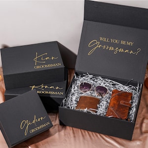Könnte beinhalten: Ein Set aus schwarzen, rechteckigen Geschenkboxen mit goldener Schrift. Eine geöffnete Box enthält silbernes Füllmaterial, braune Lederaccessoires und eine Sonnenbrille. Auf der Innenseite des Deckels steht "WILL YOU BE MY groomsman?".