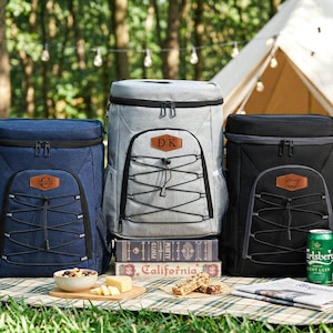 May include: Three insulated cooler backpacks in navy blue, gray, and black, with leatherette patches. The backpacks have front bungee cords and are set up for a picnic with snacks, drinks, and a tent in the background.