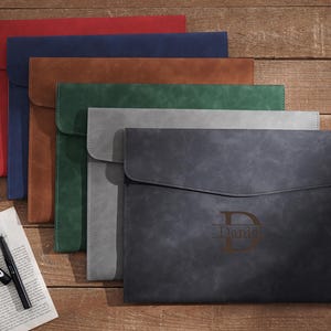 Puede incluir: Una colección de fundas de cuero para documentos en varios colores: rojo, azul marino, marrón, verde, gris y gris oscuro. El portadocumentos gris oscuro tiene grabado el nombre "Daniel" y la letra "D". Un bolígrafo y papel en primer plano.