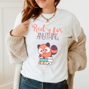 Könnte beinhalten: Weißes T-Shirt mit dem Text "Ready for Anything" in Koralle und Lila. Das Shirt zeigt einen Cartoon-Roten Panda, der auf einem Stapel Bücher sitzt. Der Panda ist orange, braun und weiß.