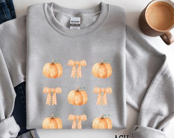 May include: A light gray sweatshirt featuring a pattern of watercolor pumpkins and orange plaid bows. The word "ASH" is printed in the bottom right corner. A brown mug of coffee is in the top right corner.