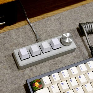 May include: A custom mechanical keyboard setup featuring a gray control panel with four white keycaps and a silver knob. A coiled gray cable connects to the panel. A portion of a keyboard with white keys and blue lettering is also visible.