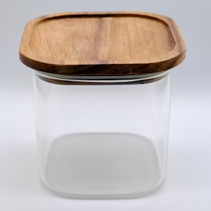 May include: A clear glass jar with a square shape and a wooden lid. The lid is a light brown color and has a square shape with rounded corners. The jar is empty and sits on a white surface.