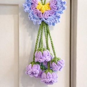 Puede incluir: Decoraci&oacute;n floral de ganchillo hecha a mano. La pieza presenta una gran flor en forma de mariposa en tonos azules, morados y amarillos, con varios capullos de flores moradas colgantes. Los tallos y el cord&oacute;n para colgar son verdes.