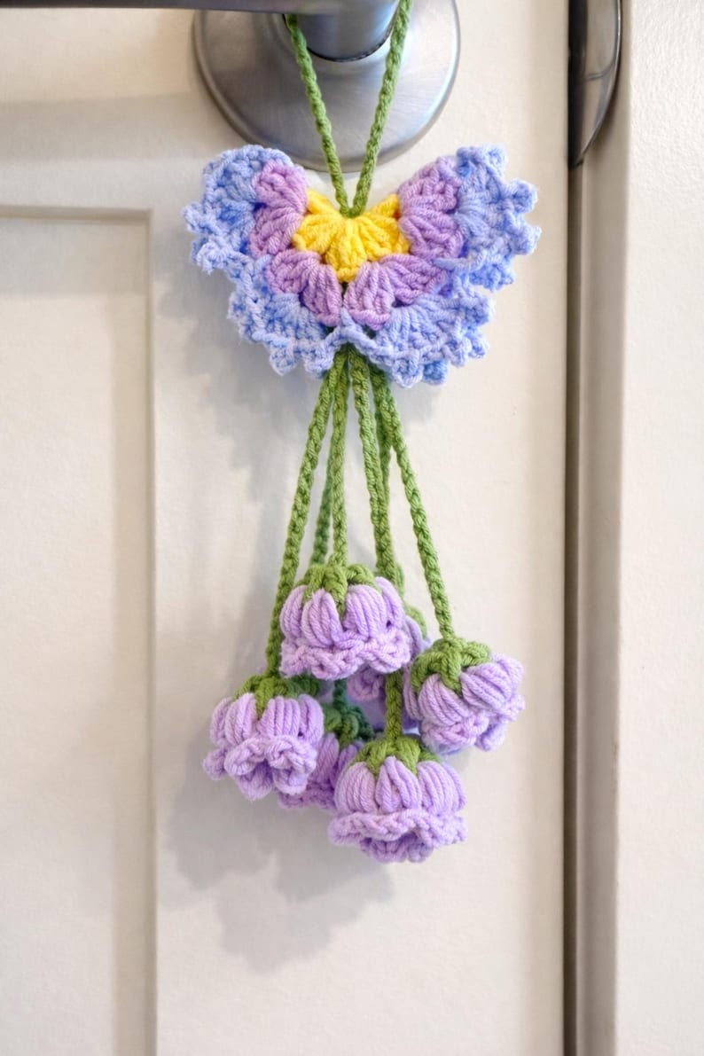 Puede incluir: Decoraci&oacute;n floral de ganchillo hecha a mano. La pieza presenta una gran flor en forma de mariposa en tonos azules, morados y amarillos, con varios capullos de flores moradas colgantes. Los tallos y el cord&oacute;n para colgar son verdes.