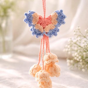 Puede incluir: Adorno de mariposa de ganchillo hecho a mano en colores pastel. La mariposa tiene secciones azules, melocot&oacute;n y rosa. Debajo de la mariposa hay tres borlas de ganchillo en forma de flor de color melocot&oacute;n, todas suspendidas por un cord&oacute;n rosa.