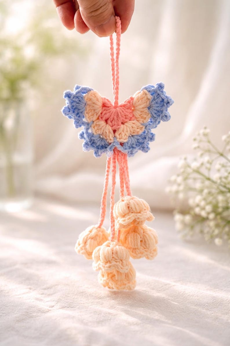 Puede incluir: Adorno de mariposa de ganchillo hecho a mano en colores pastel. La mariposa tiene secciones azules, melocot&oacute;n y rosa. Debajo de la mariposa hay tres borlas de ganchillo en forma de flor de color melocot&oacute;n, todas suspendidas por un cord&oacute;n rosa.