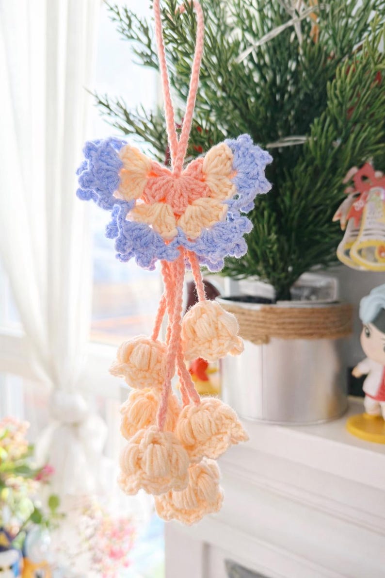 Puede incluir: Adorno de ganchillo hecho a mano con un dise&ntilde;o de mariposa en colores pastel rosa, azul claro y melocot&oacute;n. Debajo de la mariposa hay varios flecos con forma de flor de color melocot&oacute;n, todos suspendidos por una cuerda rosa. El adorno es probablemente para decoraci&oacute;n.