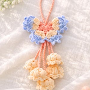 Puede incluir: Adorno de ganchillo hecho a mano en colores pastel. El adorno presenta una forma de mariposa con p&eacute;talos azules, melocot&oacute;n y crema. Debajo de la mariposa hay tres borlas en forma de flor de color crema, todas conectadas por hilo color melocot&oacute;n.