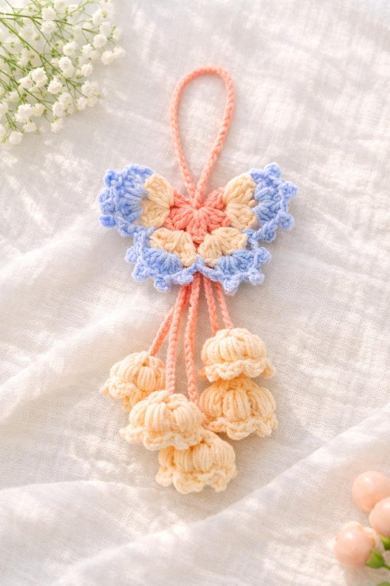 Puede incluir: Adorno de ganchillo hecho a mano en colores pastel. El adorno presenta una forma de mariposa con p&eacute;talos azules, melocot&oacute;n y crema. Debajo de la mariposa hay tres borlas en forma de flor de color crema, todas conectadas por hilo color melocot&oacute;n.