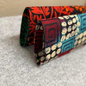 Puede incluir: Dos carteras de tela. La cartera delantera tiene un estampado de cuadrados turquesa, círculos blancos y espirales burdeos sobre un fondo negro. La cartera de atrás tiene un estampado de hojas rojas sobre fondo negro. Ambas carteras están dobladas.