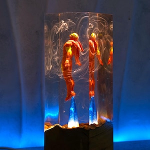 Könnte beinhalten: Eine einzigartige Lampe mit zwei orangefarbenen Figuren, die in einem transparenten, rechteckigen Block eingeschlossen sind. Die Figuren scheinen von blauen, beleuchteten Basen aufzusteigen. Die Lampe hat einen Holzsockel und einen wirbelnden, rauchigen Effekt im Inneren des transparenten Blocks.
