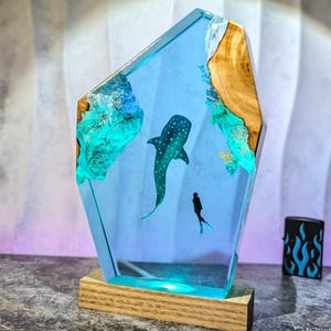 May include: A decorative resin lamp featuring a whale shark and a diver in a blue ocean scene. The lamp has a wooden base and a light blue glow. The edges are adorned with wood and turquoise accents.