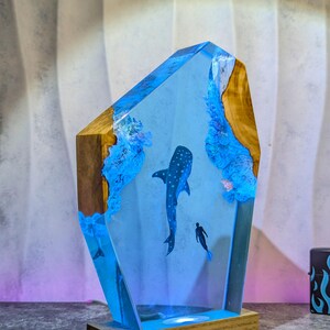 May include: A unique resin art piece featuring a whale shark and a diver in a blue ocean scene. The artwork is encased in a clear resin block with wooden edges and sits on a wooden base. The piece is illuminated with blue light.