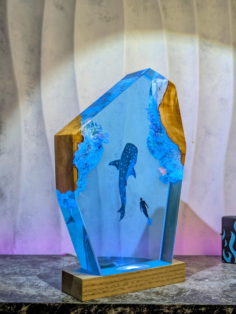 May include: A unique resin art piece featuring a whale shark and a diver in a blue ocean scene. The artwork is encased in a clear resin block with wooden edges and sits on a wooden base. The piece is illuminated with blue light.