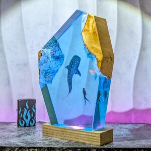 May include: A unique resin lamp featuring a whale shark and divers in a blue ocean scene. The lamp has a wooden base and a light blue resin body with a wooden accent. A black and blue flame lighter is next to the lamp.