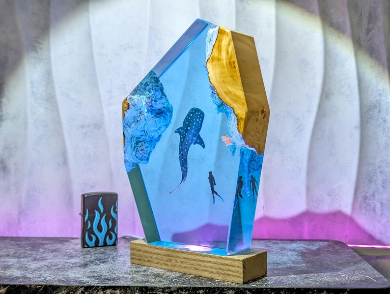 May include: A unique resin lamp featuring a whale shark and divers in a blue ocean scene. The lamp has a wooden base and a light blue resin body with a wooden accent. A black and blue flame lighter is next to the lamp.