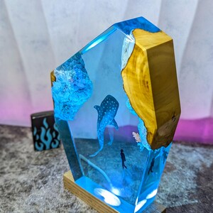 May include: A decorative resin sculpture featuring a whale shark and divers in a blue ocean scene. The sculpture has a wooden base and a geometric shape with a natural wood edge. The resin is clear, allowing the underwater scene to be visible.