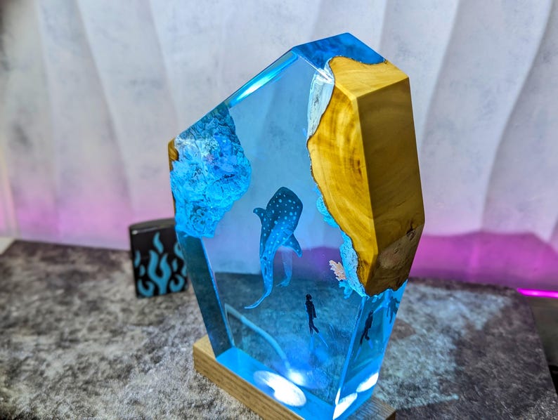 May include: A decorative resin sculpture featuring a whale shark and divers in a blue ocean scene. The sculpture has a wooden base and a geometric shape with a natural wood edge. The resin is clear, allowing the underwater scene to be visible.