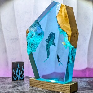 May include: A unique, handcrafted lamp featuring a whale shark and diver silhouettes encased in blue resin. The lamp has a wooden base and a section of natural wood. The design is illuminated with a soft blue light, creating a captivating underwater scene.