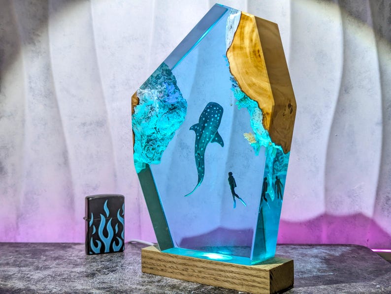 May include: A unique, handcrafted lamp featuring a whale shark and diver silhouettes encased in blue resin. The lamp has a wooden base and a section of natural wood. The design is illuminated with a soft blue light, creating a captivating underwater scene.