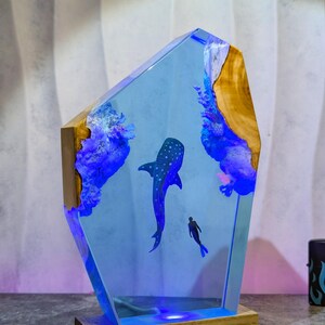 May include: A decorative resin lamp featuring a blue whale and a diver in a blue ocean scene. The lamp has a wooden base and a clear, angular resin body with embedded wood and blue accents. The lamp is lit with blue LED lights.