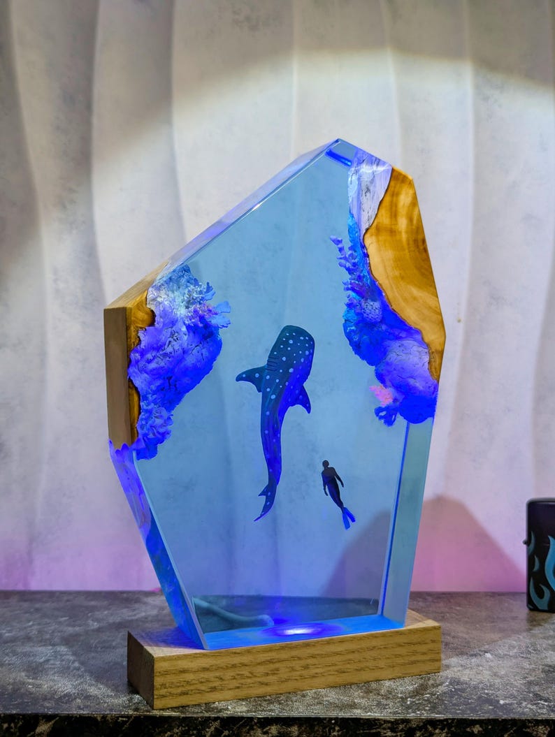 May include: A decorative resin lamp featuring a blue whale and a diver in a blue ocean scene. The lamp has a wooden base and a clear, angular resin body with embedded wood and blue accents. The lamp is lit with blue LED lights.