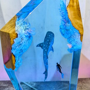 May include: A decorative resin sculpture featuring a whale shark and a diver in a blue ocean scene. The sculpture is encased in clear resin with wood accents on the sides. The whale shark is dark blue with white spots.