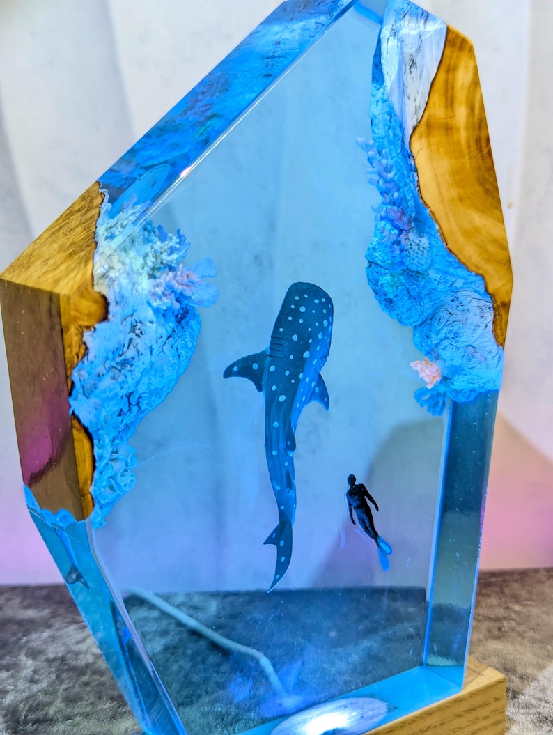 May include: A decorative resin sculpture featuring a whale shark and a diver in a blue ocean scene. The sculpture is encased in clear resin with wood accents on the sides. The whale shark is dark blue with white spots.
