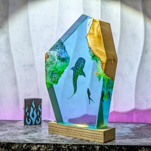 May include: A decorative resin sculpture featuring a whale shark and divers in a blue ocean scene. The sculpture has a wooden base and a light blue and green color scheme. A black lighter with blue flames is on the left.