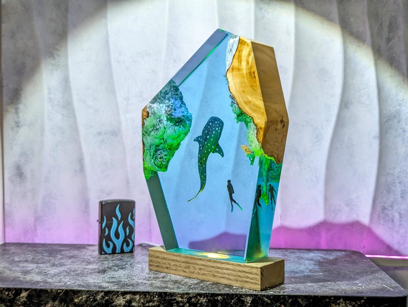 May include: A decorative resin sculpture featuring a whale shark and divers in a blue ocean scene. The sculpture has a wooden base and a light blue and green color scheme. A black lighter with blue flames is on the left.
