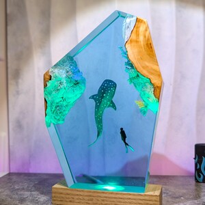 May include: A decorative resin sculpture featuring a whale shark and a diver in a blue ocean scene. The sculpture has a wooden base and a light that illuminates the scene. The edges are a light blue color, and the sculpture has a wooden accent.