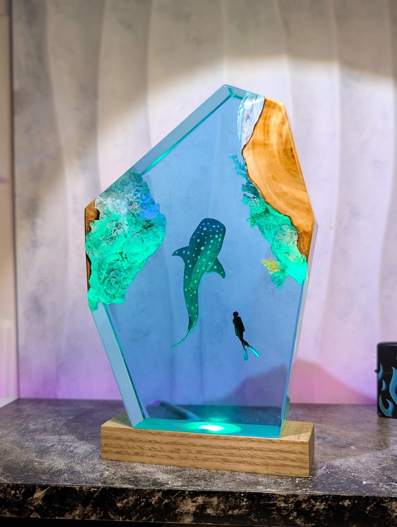 May include: A decorative resin sculpture featuring a whale shark and a diver in a blue ocean scene. The sculpture has a wooden base and a light that illuminates the scene. The edges are a light blue color, and the sculpture has a wooden accent.