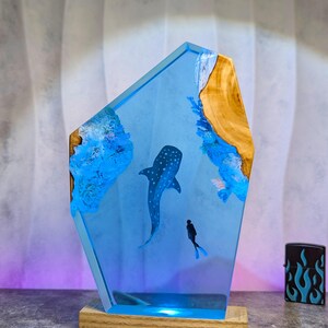 May include: A blue resin lamp featuring a whale shark and a diver. The lamp has a wooden base and a light blue glow. The design includes blue coral and wood accents, creating an underwater scene.
