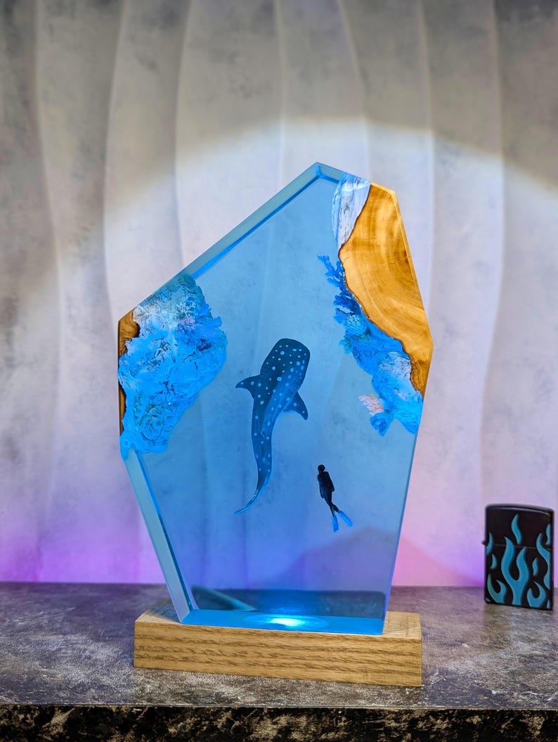 May include: A blue resin lamp featuring a whale shark and a diver. The lamp has a wooden base and a light blue glow. The design includes blue coral and wood accents, creating an underwater scene.