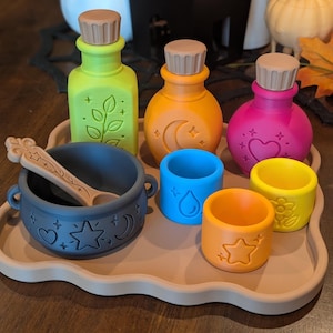 Peut inclure: Un ensemble de flacons de potion colorés, de petits gobelets et un chaudron gris foncé avec une cuillère assortie, disposés sur un plateau beige. Les objets présentent des motifs en relief incluant des étoiles, lunes, cœurs, fleurs et gouttes d'eau.