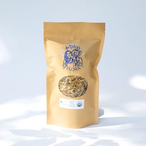 May include: A brown paper bag of dried sunflower petals with a clear oval window. The bag features a blue logo with the words "drittless FLORA" and a small green and white organic label. The bag is filled with yellow and brown petals.