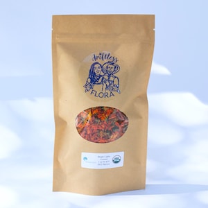 May include: A brown paper bag with a clear oval window displaying dried orange flowers. The bag has a blue logo that says "driftless flora" and a white label with text. The bag contains 1 oz of Bright Lights Calendula.