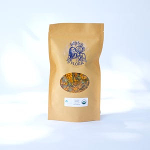 May include: A brown paper bag with a clear oval window displaying dried orange calendula flowers. The bag has a blue logo with two figures and the text "drifltess FLORA". A white label with text and an organic seal is visible.