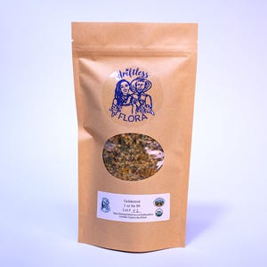 May include: A brown paper bag with a clear oval window displaying dried goldenrod. The bag features a blue and white logo with the text "driftless flora" and a label with product information and an organic certification.