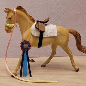 May include: A tan-colored toy horse with a brown saddle, a white and gray saddle blanket, and a blue, red, and gold award ribbon. The horse has a pink and yellow lead rope. The horse is a toy.