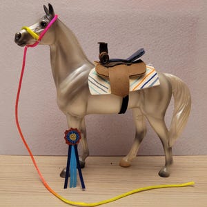 May include: A gray toy horse with a saddle, bridle, and a colorful ribbon. The saddle is brown with a blue and white striped pad. The bridle is pink and yellow, and the ribbon is blue, orange, and yellow.