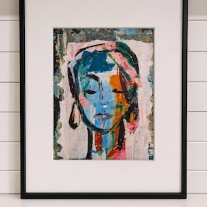May include: A framed abstract portrait painting featuring a stylized face rendered in bold, expressive brushstrokes of blue, orange, pink, and black. The artwork is set within a white mat and a thin black frame, showcasing a textured, layered paint application.