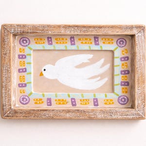 May include: A framed painting of a white dove in flight. The artwork is set within a light brown wooden frame. The inner border is decorated with a repeating pattern of purple, yellow, and blue shapes.