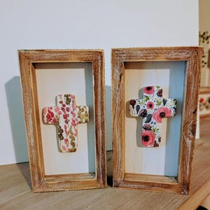 Puede incluir: Dos cruces enmarcadas con motivos florales. Las cruces están sobre un fondo blanco dentro de marcos de madera marrón claro y rústico. Una cruz presenta flores rosas y verdes, la otra rosas rosas.