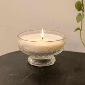 May include: A burning white candle in a clear, textured glass bowl. The candle is on a dark surface, with a plant in the background. The candle flame is bright against the soft, warm glow of the wax.