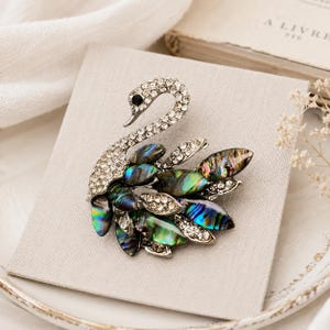 May include: Swan brooch with a silver-tone body encrusted with clear crystals. The brooch features iridescent abalone shell feathers in shades of green, blue, and purple. A black crystal forms the eye.