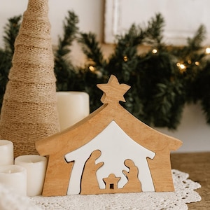 May include: Wooden nativity scene with a star on top, featuring a white interior and silhouettes of Mary, Joseph, and baby Jesus. A decorative Christmas tree and candles are in the background. The scene is set on a white lace doily.