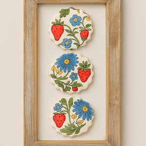 May include: Three round ceramic coasters with a floral and strawberry design, framed in a light brown wooden frame. Each coaster features a white background with red strawberries, blue flowers, and green leaves.